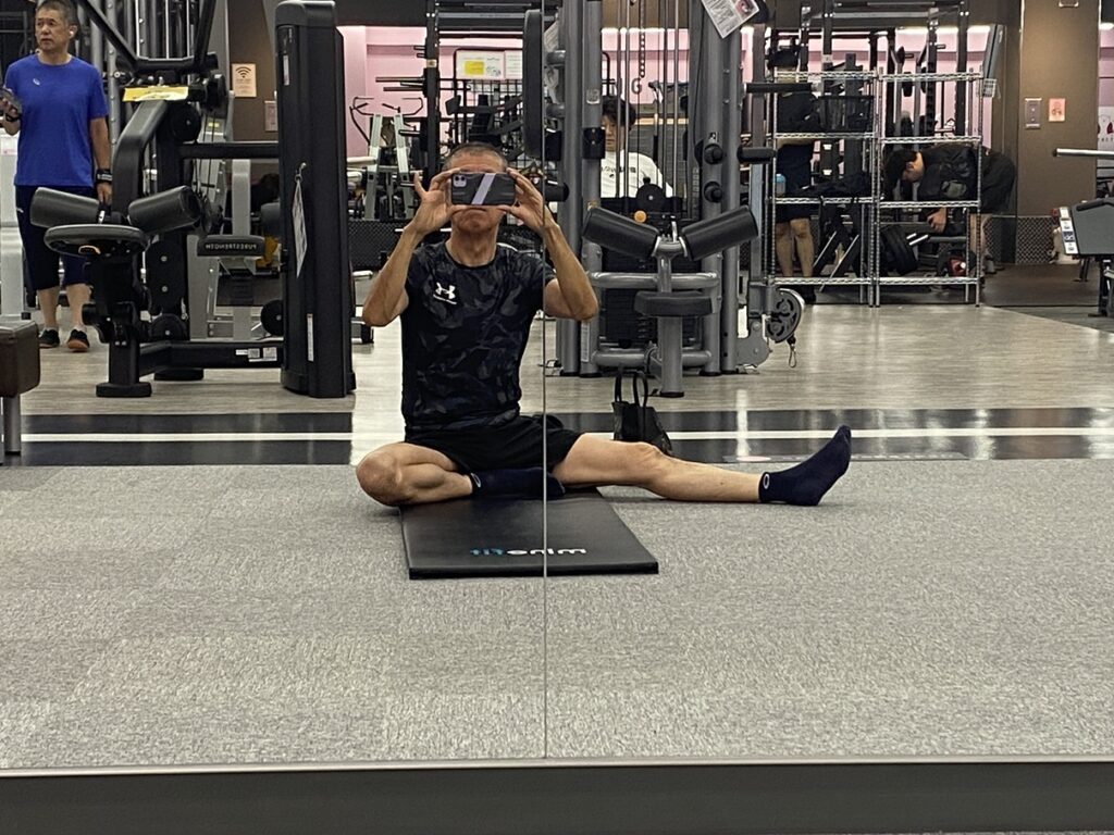 スポーツジムのストレッチゾーンで、黒いスポーツウェア姿の男性がマットの上に座り、片足を伸ばしてスマートフォンで自撮りしている様子。背景には筋トレマシンやトレーニング中の利用者が見える。」