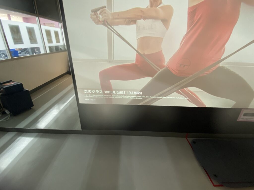 スポーツジムのスタジオで、壁のスクリーンに次のクラス『VIRTUAL DANCE（45分）』の案内が映し出されている様子。手前の床にマットが置かれ、レッスン開始を待つ静かな雰囲気。
