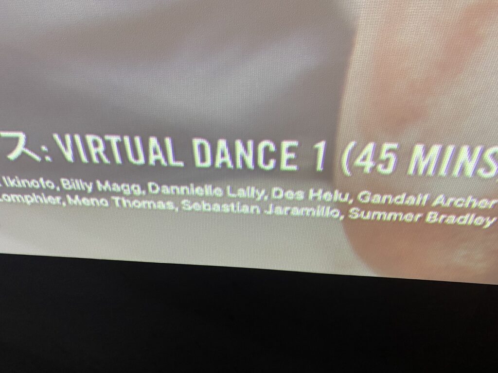 ジムのスタジオスクリーンに『LesMills Virtual Dance 1（45分）』と表示されたリリースNo.1のプログラム案内。インストラクターの名前一覧も画面下に映し出されている。