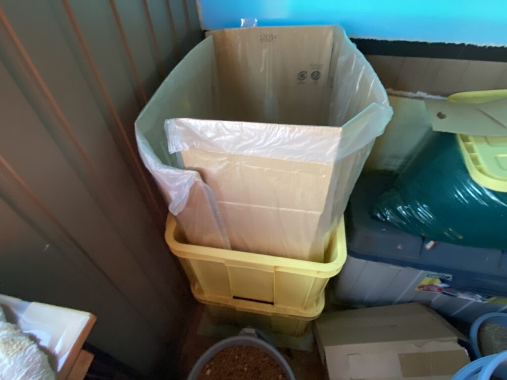 段ボールとごみ袋をセットした状態の落ち葉用ボックスコンテナ。周囲の資材や収納箱と並び、作業スペースが整理されている