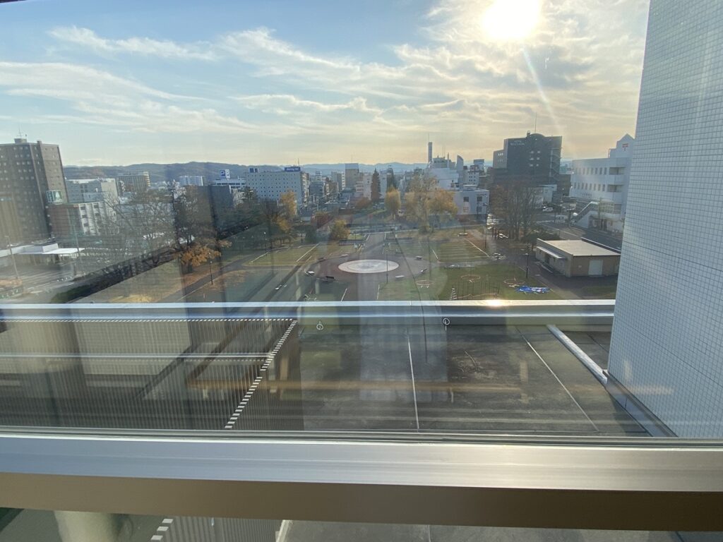 病室の窓から見下ろした北見市中心部の街並み。晴れた空の下、公園の遊具や木々、遠くの建物が見え、午後の日差しがやわらかく差し込んでいる。