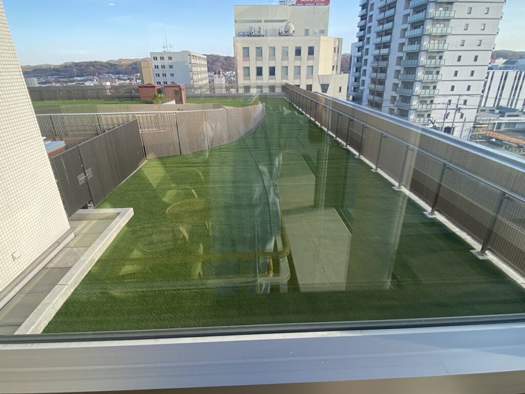 建物の屋上にある人工芝の庭園。手すりに囲まれた明るい空間で、遠くに低い山並みと市街地の建物が見える。
