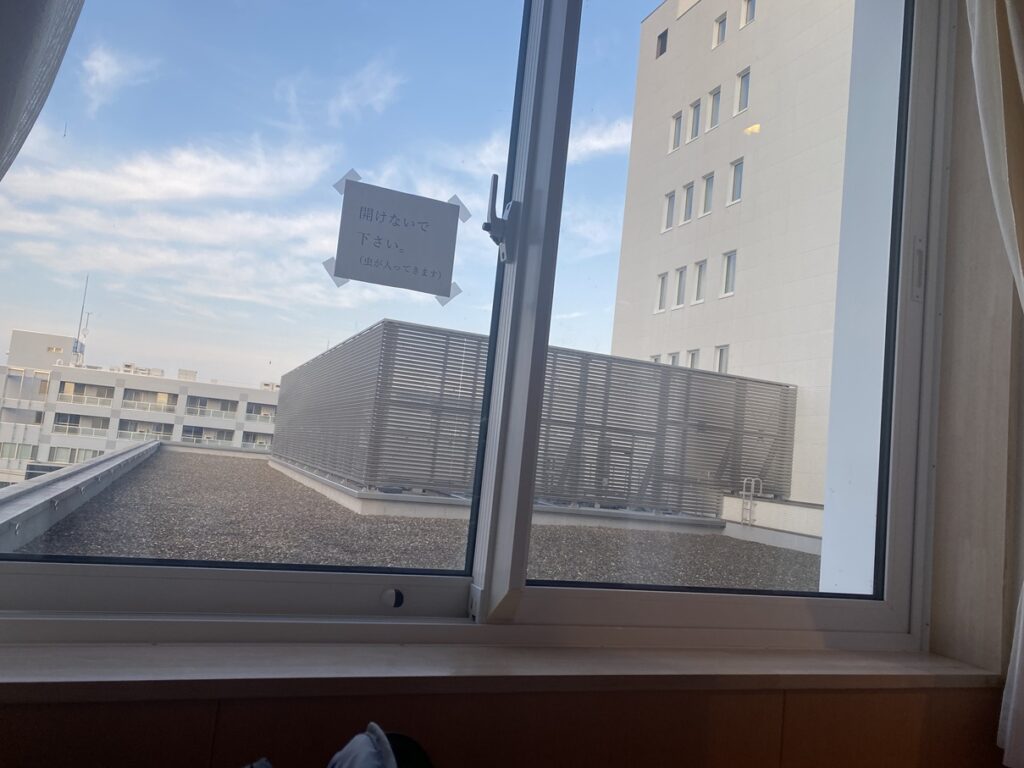 病室の窓から見える外の風景。向かいの建物と青空が見え、窓ガラスには「開けないでください(虫が入ってきます)」という貼り紙がある。