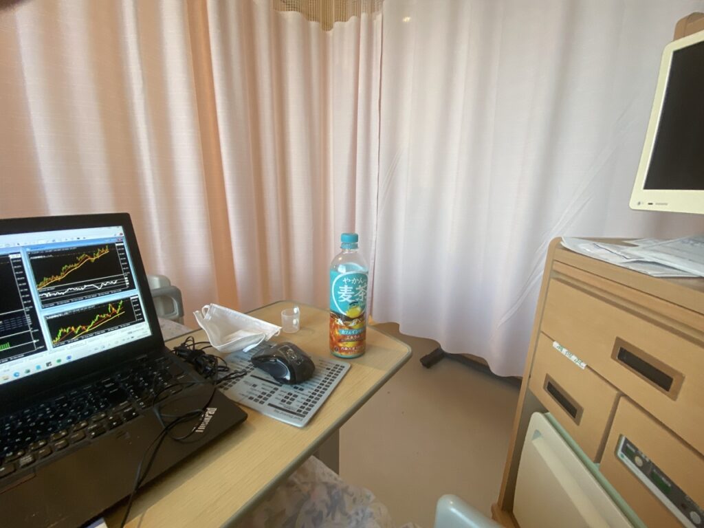 病室のベッド横にある小さな机の上にノートパソコンとマウス、マスク、そして「やかんの麦茶」が置かれている。パソコン画面にはトレード用のチャートが複数表示されている。