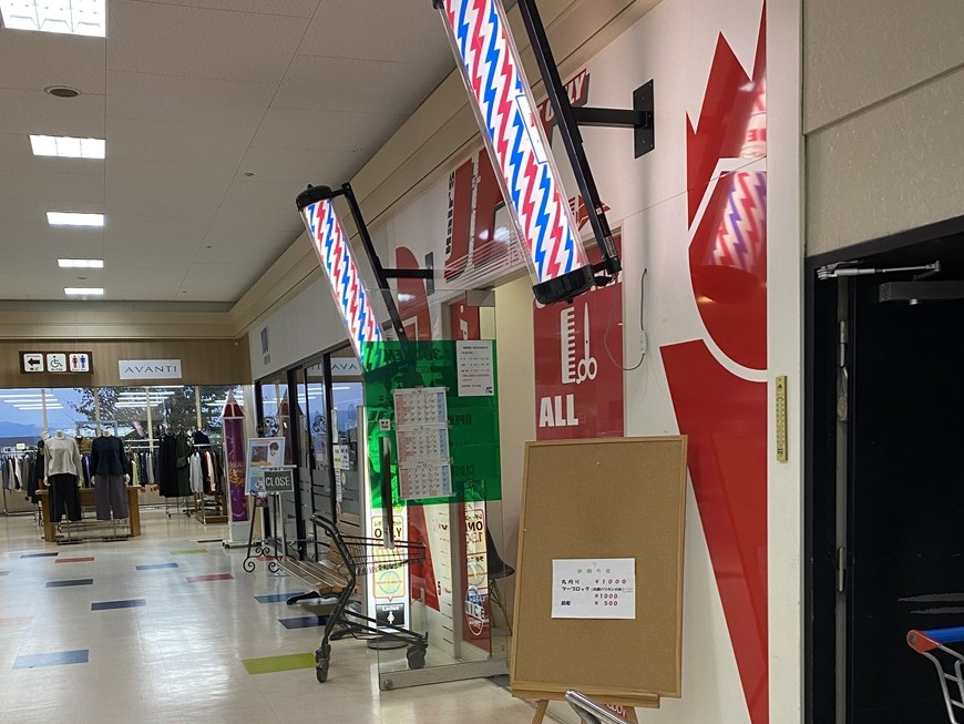 ショッピングモール内の理容室入り口。赤・青・白のサインポールが2本並び、理髪店の目印が光る床屋の外観。料金表や案内板が設置され、清潔感のある店構えが印象的。カット料金や営業時間が掲示されており、買い物ついでに立ち寄れる便利なヘアカット専門店の様子。