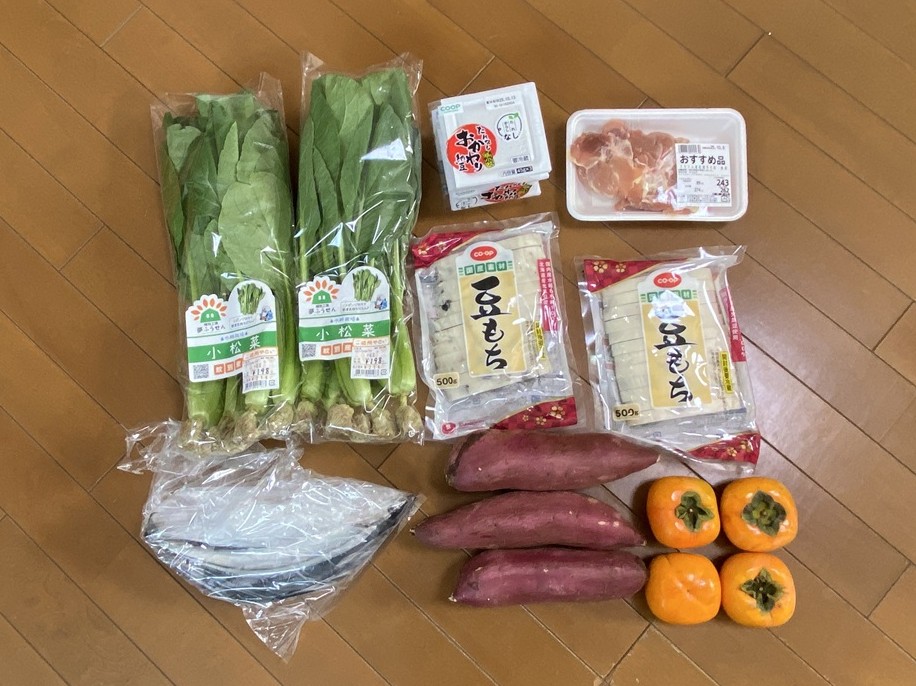 今日の買い物で購入した食材を床に並べた写真。小松菜3束、納豆、鶏もも肉、豆もち2袋、サツマイモ2本、柿4個、そしてサンマ6尾。秋の旬食材を中心に、主食・たんぱく質・野菜・果物をバランスよく揃えた健康的な買い物内容。