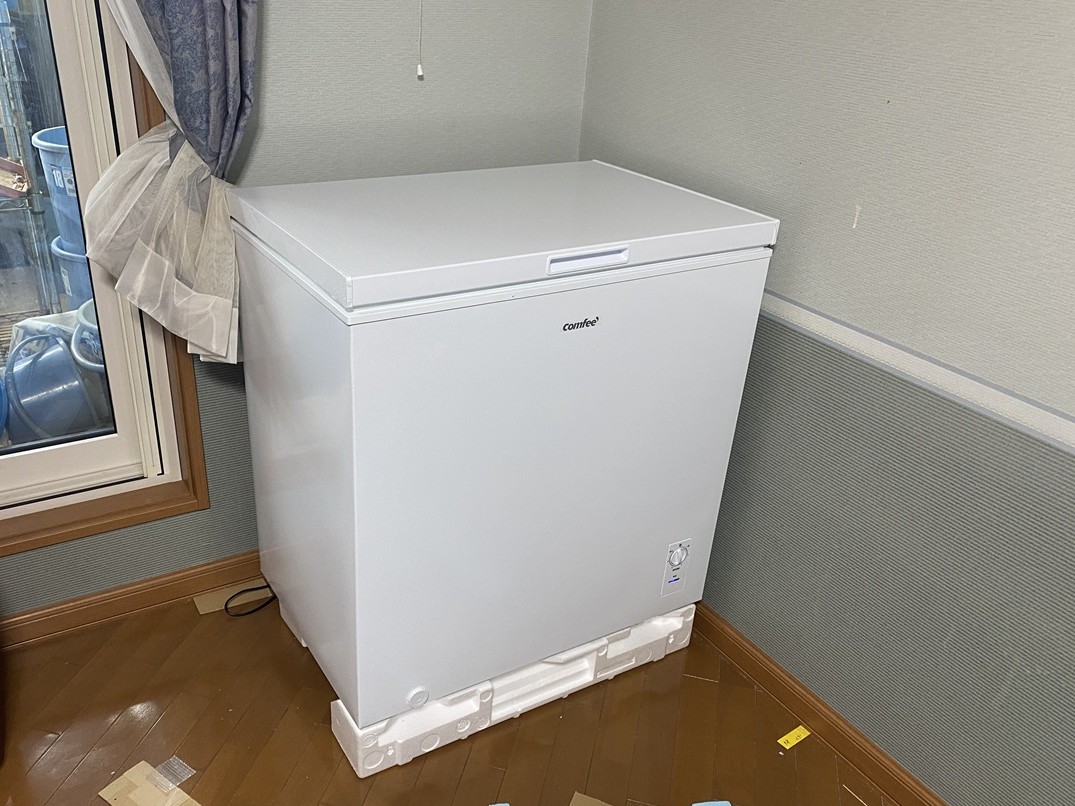 新しく購入した白い上開き冷凍庫（Comfee製）を寝室の角に設置した様子。床には保護用の発泡スチロールが残り、設置直後の清潔感ある状態がわかる。