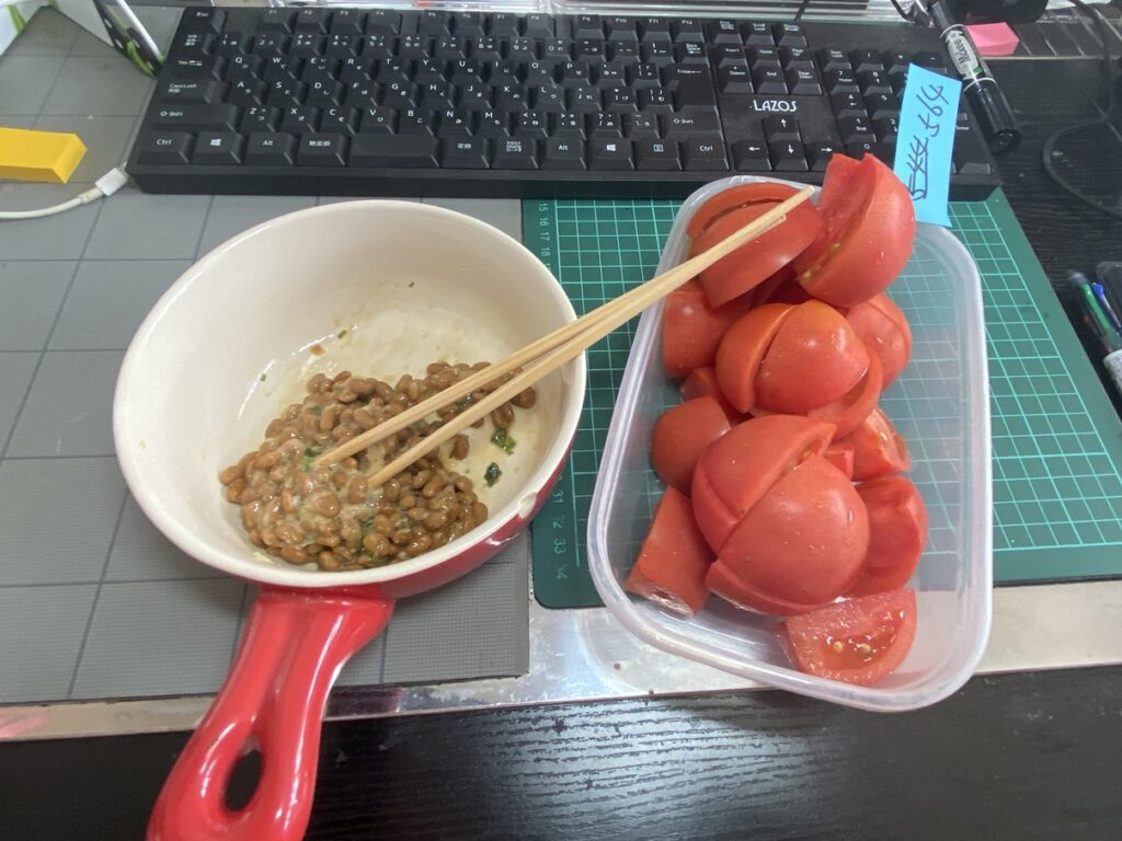 器に入った納豆と、タッパーに盛られたカットトマトを箸と一緒に並べた間食の様子。