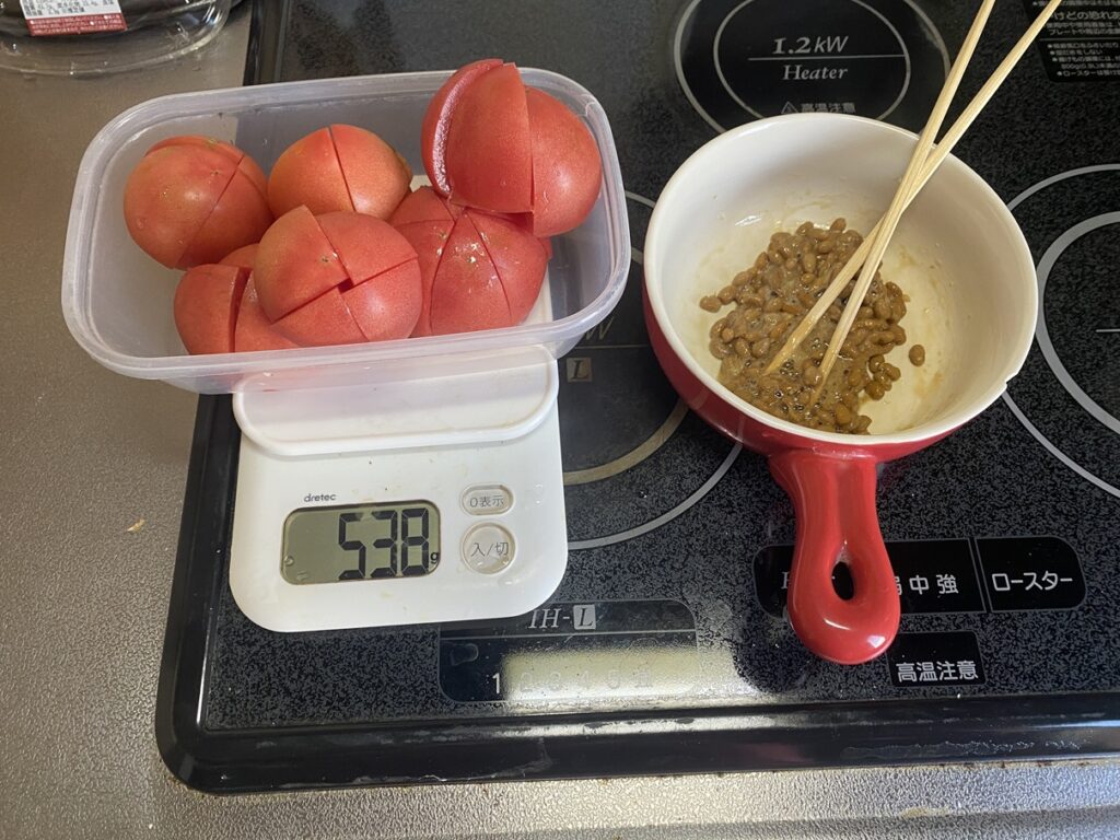 トマト538gを計量した容器と、赤い小鍋に入れた納豆を並べた健康食の準備。トマトのリコピンやビタミンC、納豆のたんぱく質やナットウキナーゼを組み合わせた栄養バランス食で、ダイエットや血流改善、免疫力アップに効果的なヘルシーメニュー。