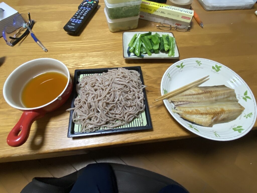 晩ご飯のメニューは、香ばしく焼いたホッケの開き、ゆでた小松菜、そして風味豊かな十割そば。 赤い鍋のつゆにそばをつけて食べるスタイルで、脂質を抑えつつたんぱく質とミネラルをしっかり摂取できる和食の健康献立。 ダイエット中にもおすすめの低カロリーで栄養バランスの良い夕食。