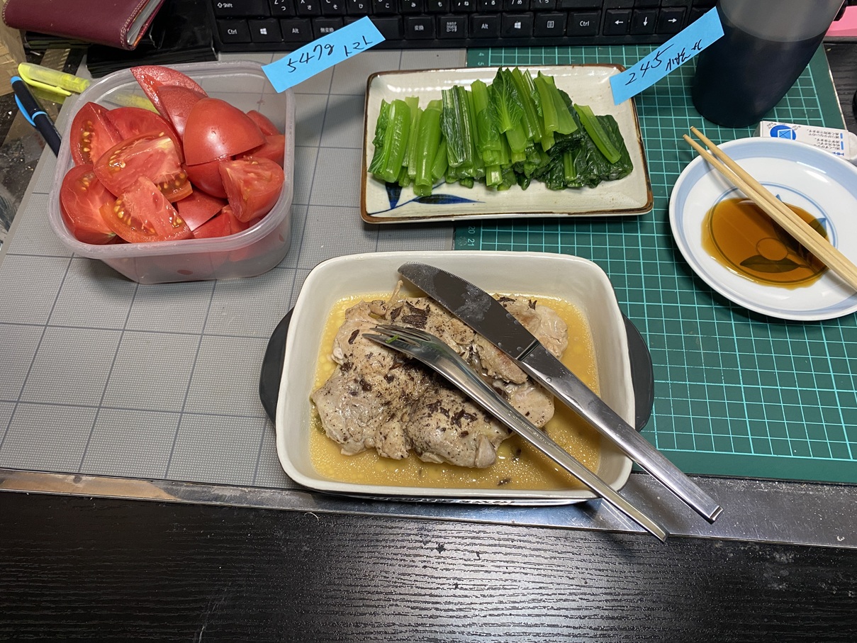 作業デスクの上に並べられた夕食。容器いっぱいのカットトマト、湯がいた小松菜のおひたし、ソースに浸った鶏むね肉のソテー、しょうゆ皿と箸、飲み物の入ったコップが並んでいる。
