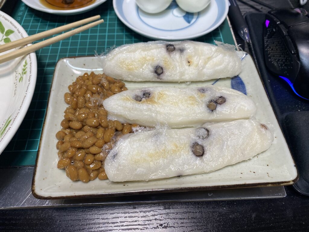 皿の上に黒豆入りの焼き餅が3本並び、隣には粘り気のある納豆が添えられている。和の朝食らしい素朴な組み合わせで、湯気の立つ餅が食欲をそそる。