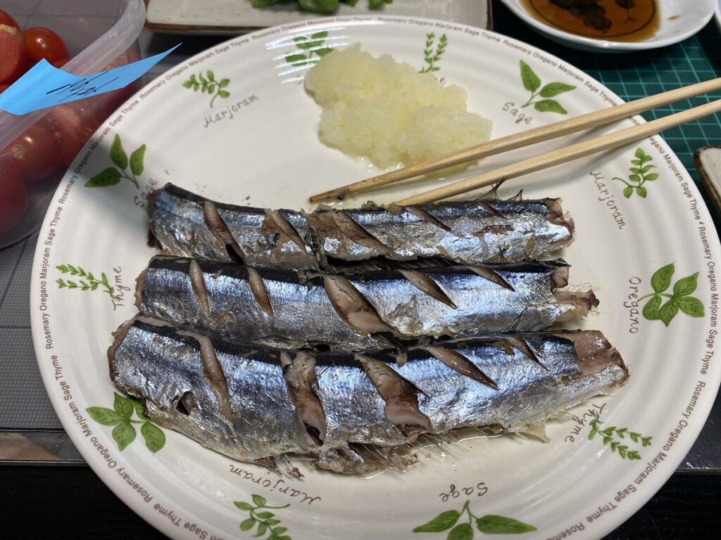 焼きサンマ4尾が皿に並び、切れ込みから脂がにじむ。右上には大根おろしが添えられ、横には箸が置かれている。秋の味覚らしい、シンプルで香ばしい和食の一皿。