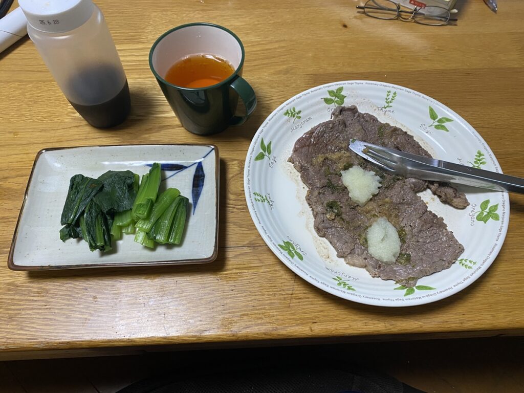 湯がいた小松菜のおひたしと、大根おろしを添えた牛肩ロースステーキの定食。緑のマグカップにはルイボスティーが注がれ、木製テーブルの上に並んだ、シンプルで健康的な夕食の風景。