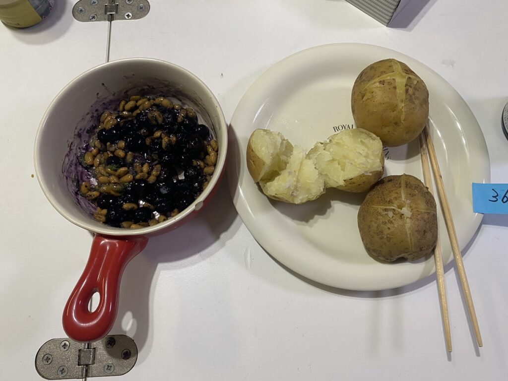 赤い小鍋の納豆とブルーベリー、隣の皿に蒸したじゃがいも3個(1個は割って中身が見える)、割り箸付き。