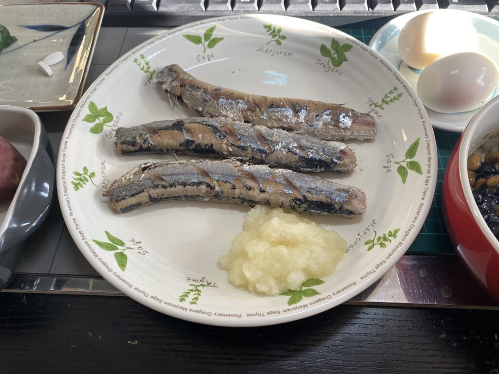 白い丸皿に焼いたサンマが3本並び、手前にたっぷりの大根おろし。周囲に半熟卵や納豆ブルーベリー、さつまいも、小皿が配置されている。