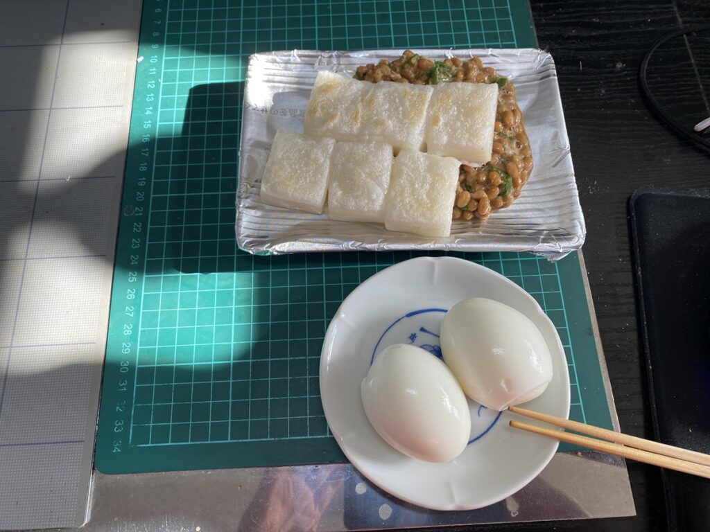 焼いた切り餅を納豆の上にのせた「納豆もち」と、ゆで卵2個を並べた昼食。
緑のカッティングマットの上に置かれ、シンプルながらも栄養バランスのとれた一食。
